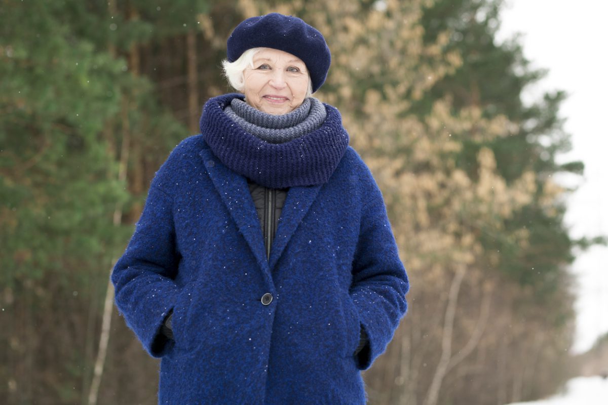 Waist up portrait of modern senior woman posing in winter forest and smiling at camera