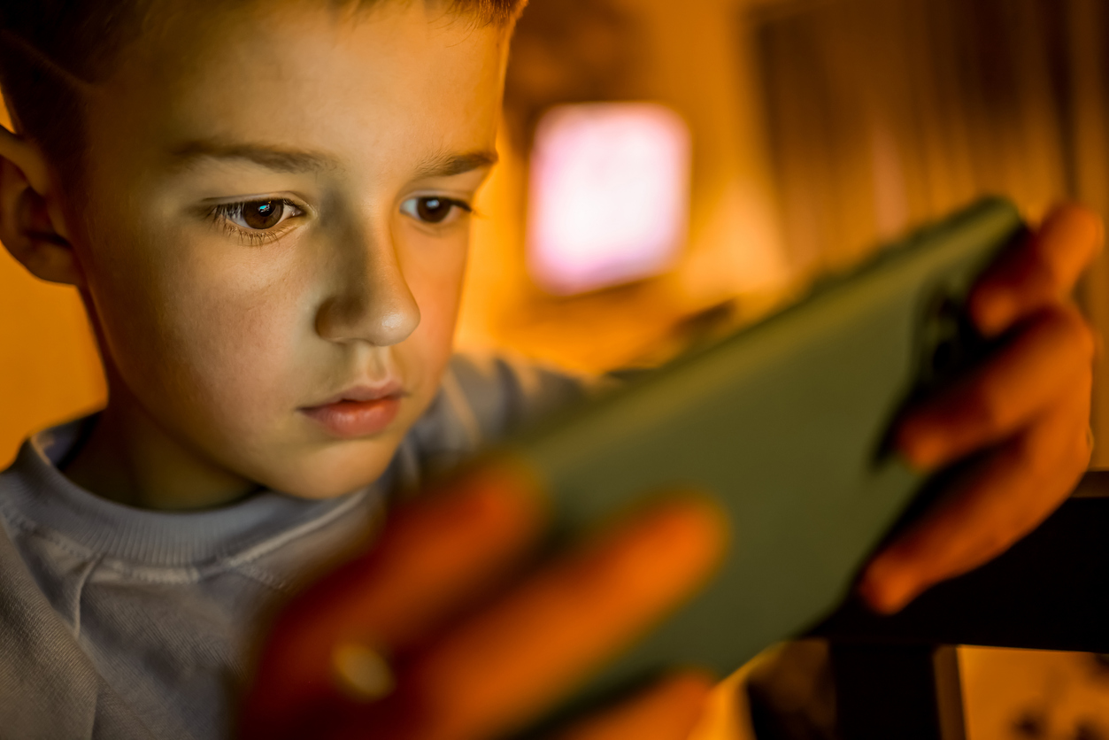 A teenage boy sits alone, illuminated by the glow of a smartphone screen, symbolizing the impact of excessive screen time on adolescent mental health